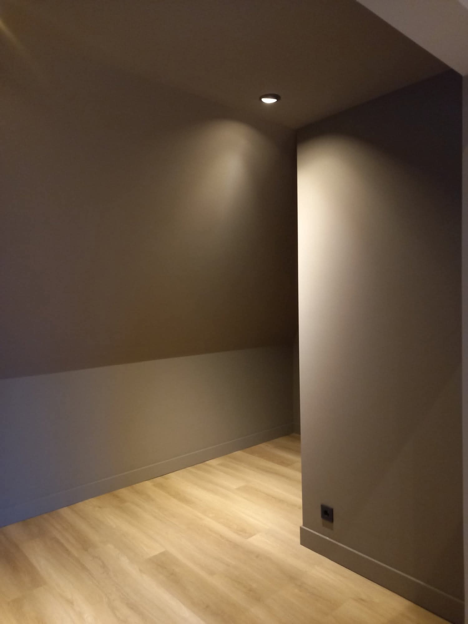 Pokój na poddaszu po remoncie: szare ściany, drewniana podłoga, oświetlenie punktowe. Widoczny fragment skosu i listwa przypodłogowa w kolorze ścian.