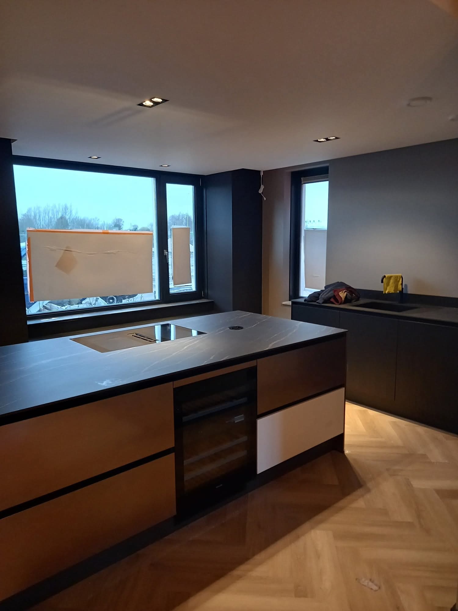 Nowoczesna kuchnia z wyspą i parkietem w jodełkę w Rzeszowie. Okno z widokiem na zewnątrz, minimalistyczne oświetlenie sufitowe. Eleganckie wykończenie.