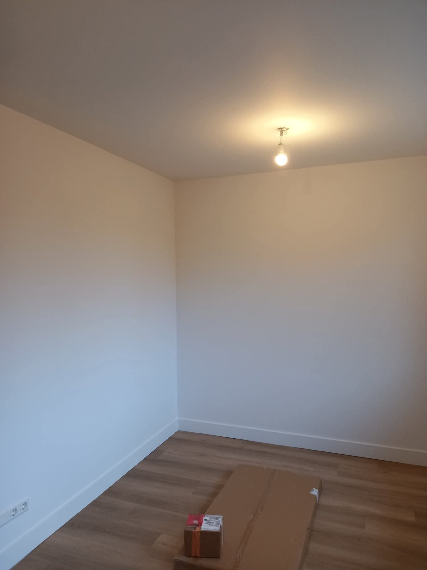 Pomalowane na jasno pomieszczenie z panelami podłogowymi i listwami przypodłogowymi. Na podłodze leżą kartony. Widoczna żarówka na suficie.