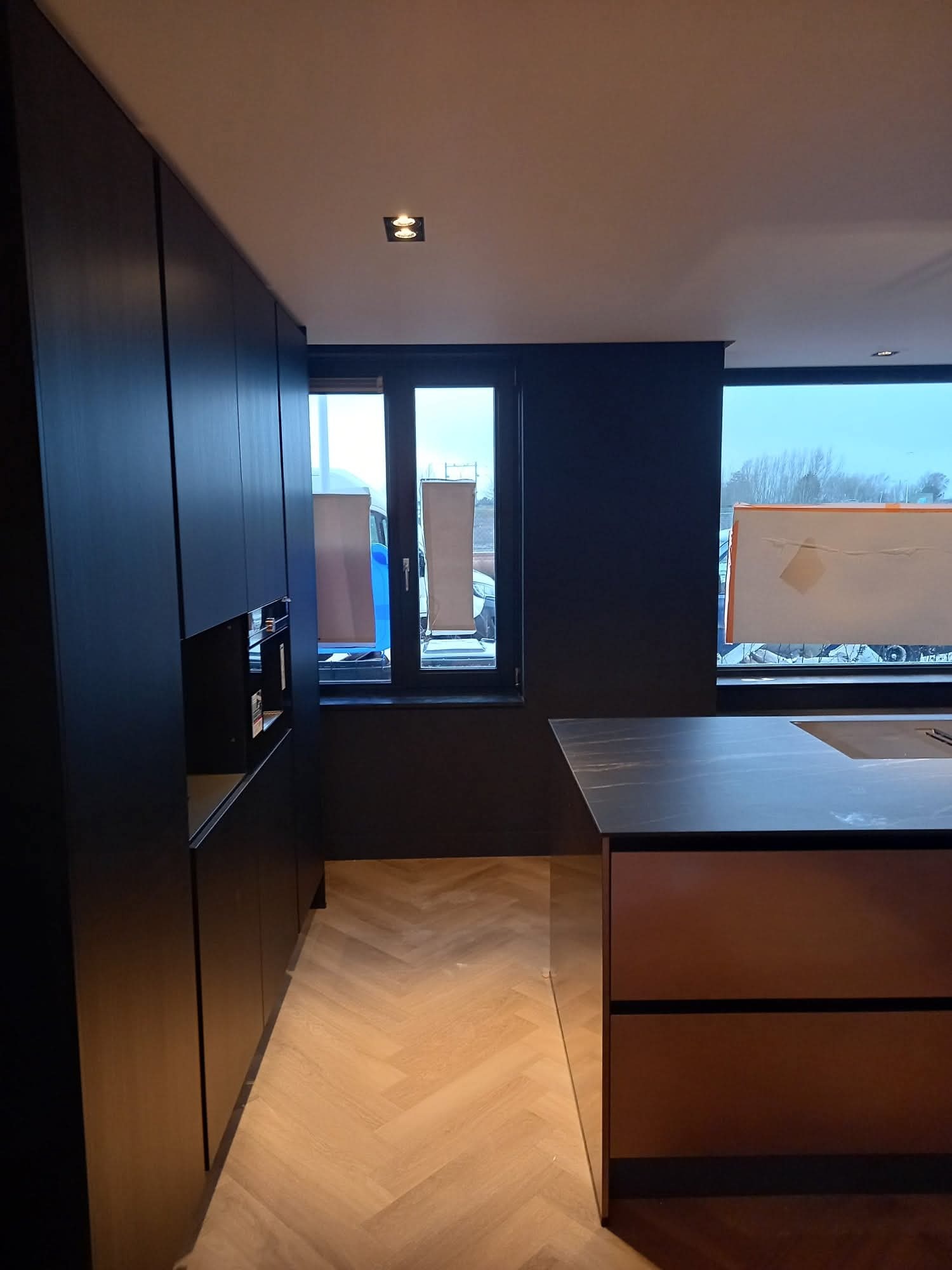 Nowoczesna kuchnia z ciemnymi szafkami i drewnianą podłogą w jodełkę. Widoczne okna z widokiem na zewnątrz i wyspa kuchenna z blatem imitującym kamień.