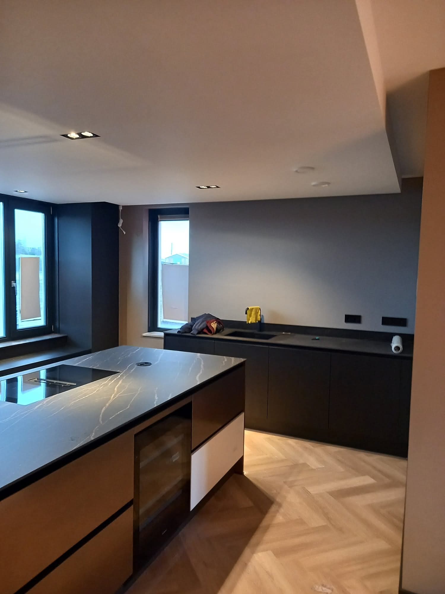 Nowoczesna kuchnia z wyspą i parkietem w jodełkę. Ciemne szafki kontrastują z jasnym blatem. Widoczne okna i oświetlenie sufitowe.