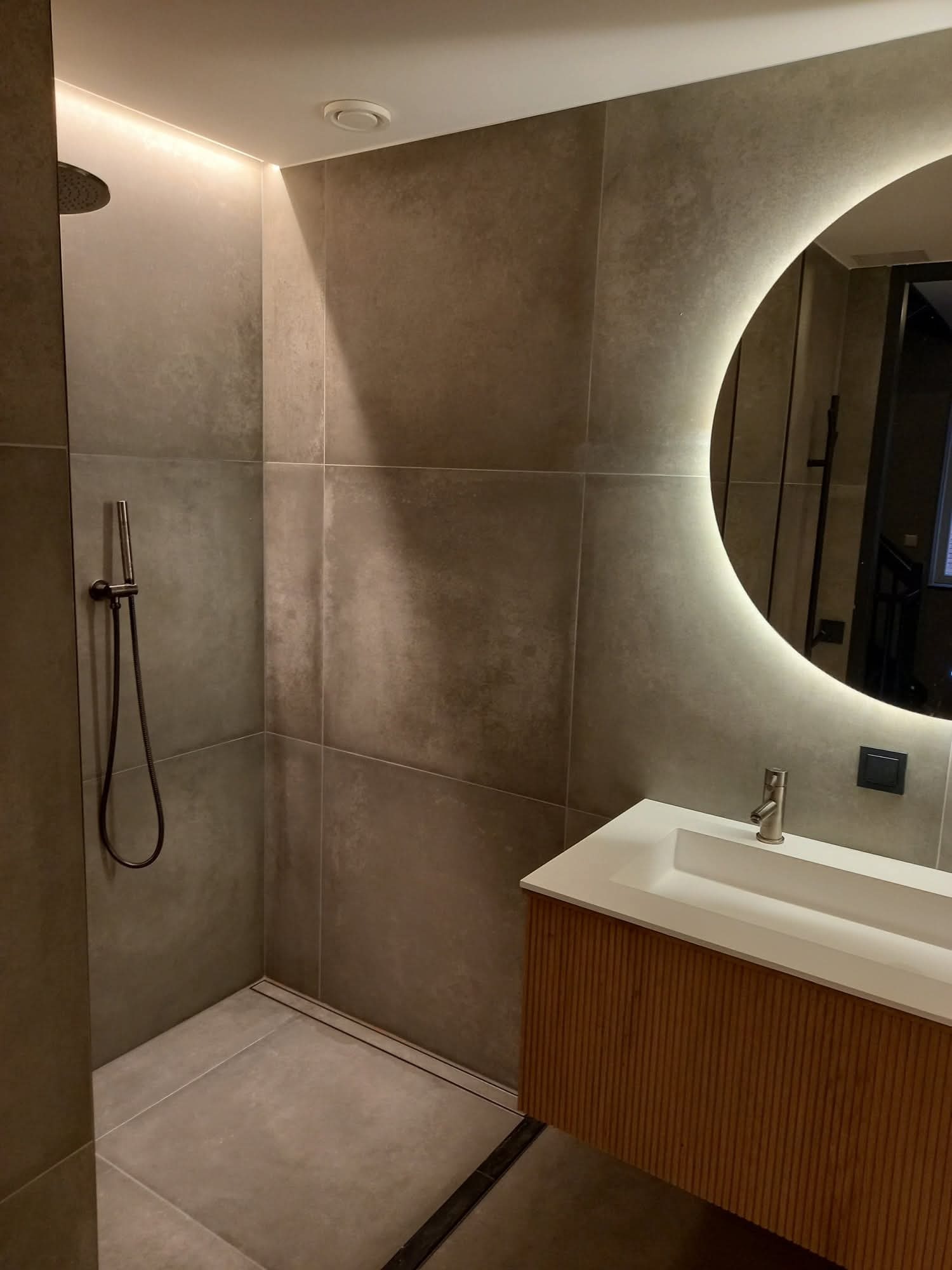 Nowoczesna łazienka z szarymi płytkami, kabiną prysznicową typu walk-in i okrągłym lustrem z podświetleniem LED. Umywalka nablatowa z drewnianą szafką.