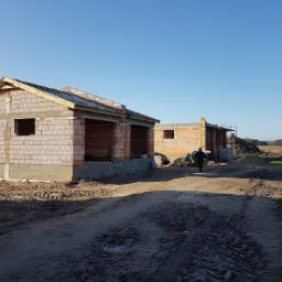 Budowa dwóch domów jednorodzinnych w stanie surowym otwartym, z widocznymi ścianami z bloczków ceramicznych, drewnianą konstrukcją dachu i osobą w oddali, na tle błękitnego nieba.