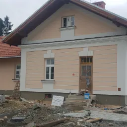 Dom w Nowym Targu z jasną elewacją w trakcie prac wykończeniowych, widoczne materiały budowlane i pracownicy układający kostkę brukową przed budynkiem.