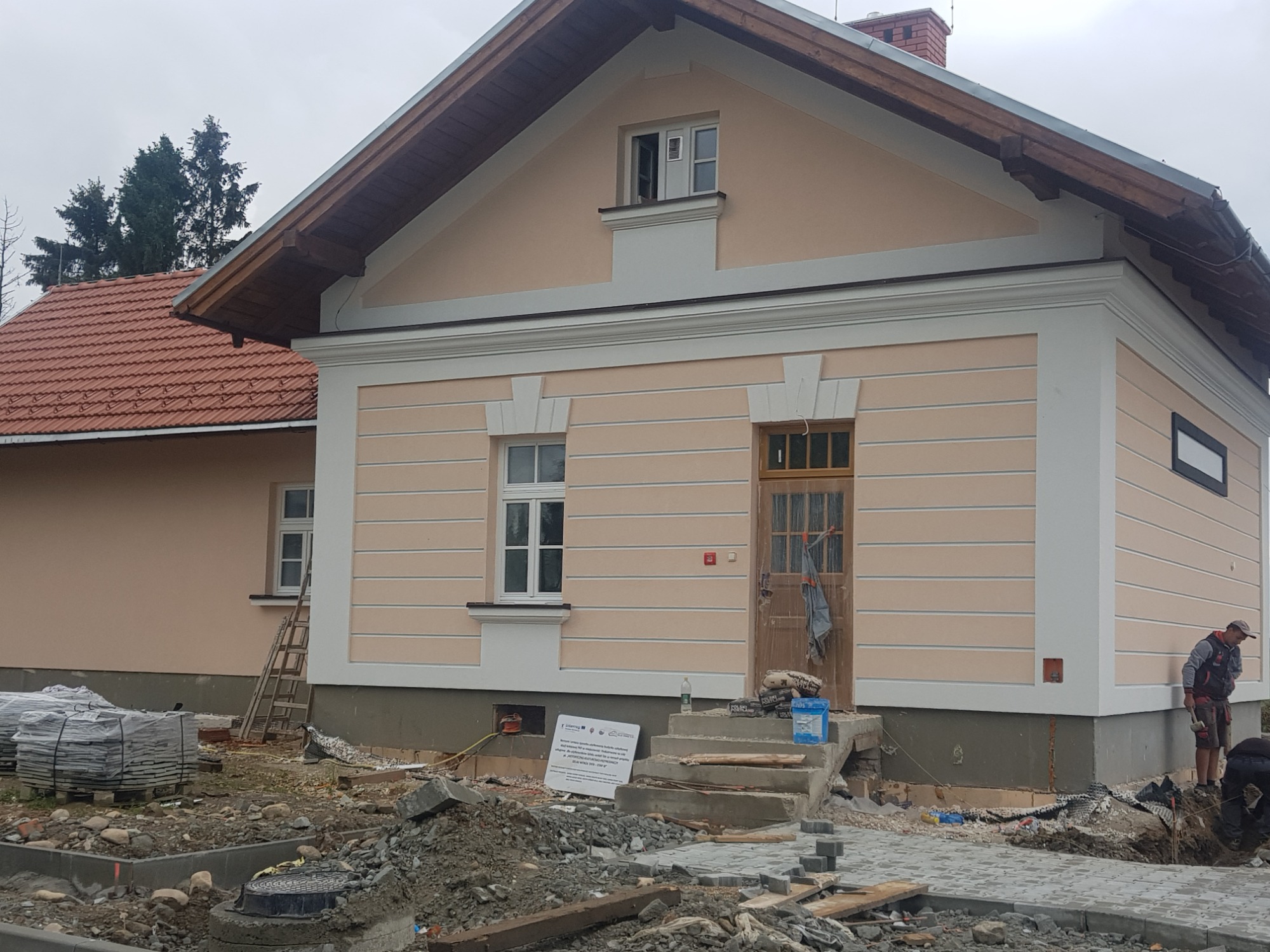 Dom w Nowym Targu z jasną elewacją w trakcie prac wykończeniowych, widoczne materiały budowlane i pracownicy układający kostkę brukową przed budynkiem.