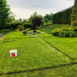 Przygotowanie terenu pod trawnik z rolki: wyznaczone linie cięcia w trawie, widoczny kubeł z logo BauMit, taczka z ziemią i rozłożone fragmenty darni na zielonym trawniku w ogrodzie.