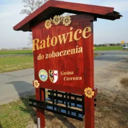Drewniany szyld informacyjny z nazwą miejscowości Ratowice i herbem Gminy Czernica, ozdobiony kwiatami, umieszczony przy drodze.