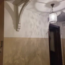 Eleganckie wykończenie wnętrza z dekoracyjną ścianą w odcieniach beżu i bieli, ozdobnymi wspornikami pod sufitem i klasyczną lampą wiszącą z wzorzystym kloszem, widoczne fragmenty stolarki drzwiowej.