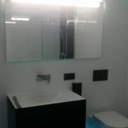 Nowoczesna łazienka z podświetleniem LED: podwieszana szafka umywalkowa z białą umywalką, lustro z oświetleniem, wisząca toaleta i czarne płytki podłogowe z efektem połysku.