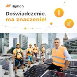 Autoryzowany partner Hymon Fotowoltaika i pompy ciepła - Zespół instalatorów fotowoltaiki na dachu budynku z panoramą miasta w tle, lider zespołu w pomarańczowej kamizelce odblaskowej stoi na pierwszym planie, logo firmy Hyman w lewym górnym rogu.
