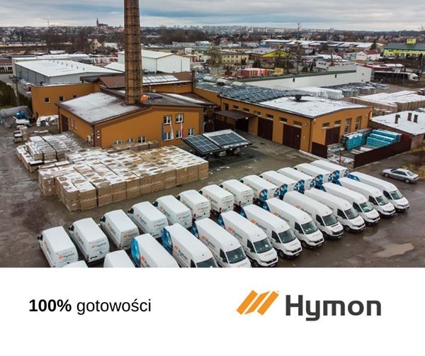 Widok z góry na teren firmy z panelami słonecznymi na dachu, stosem zapakowanych materiałów i flotą białych samochodów dostawczych z logo firmy.