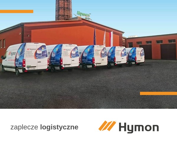 Flota białych samochodów dostawczych z logo firmy Hymen zaparkowana przed budynkiem z cegły i kominem, widoczne flagi Polski i Unii Europejskiej.