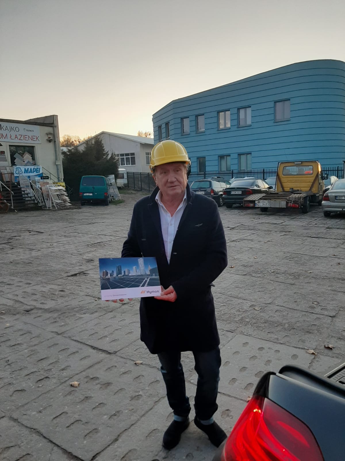 Mężczyzna w kasku budowlanym prezentuje broszurę z wizualizacją paneli fotowoltaicznych na tle miejskiego krajobrazu, stojąc na placu budowy z budynkami w tle.