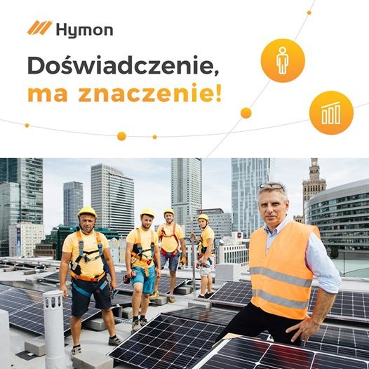 Zespół instalatorów fotowoltaiki na dachu budynku z panoramą miasta w tle, lider zespołu w pomarańczowej kamizelce odblaskowej stoi na pierwszym planie, logo firmy Hyman w lewym górnym rogu.