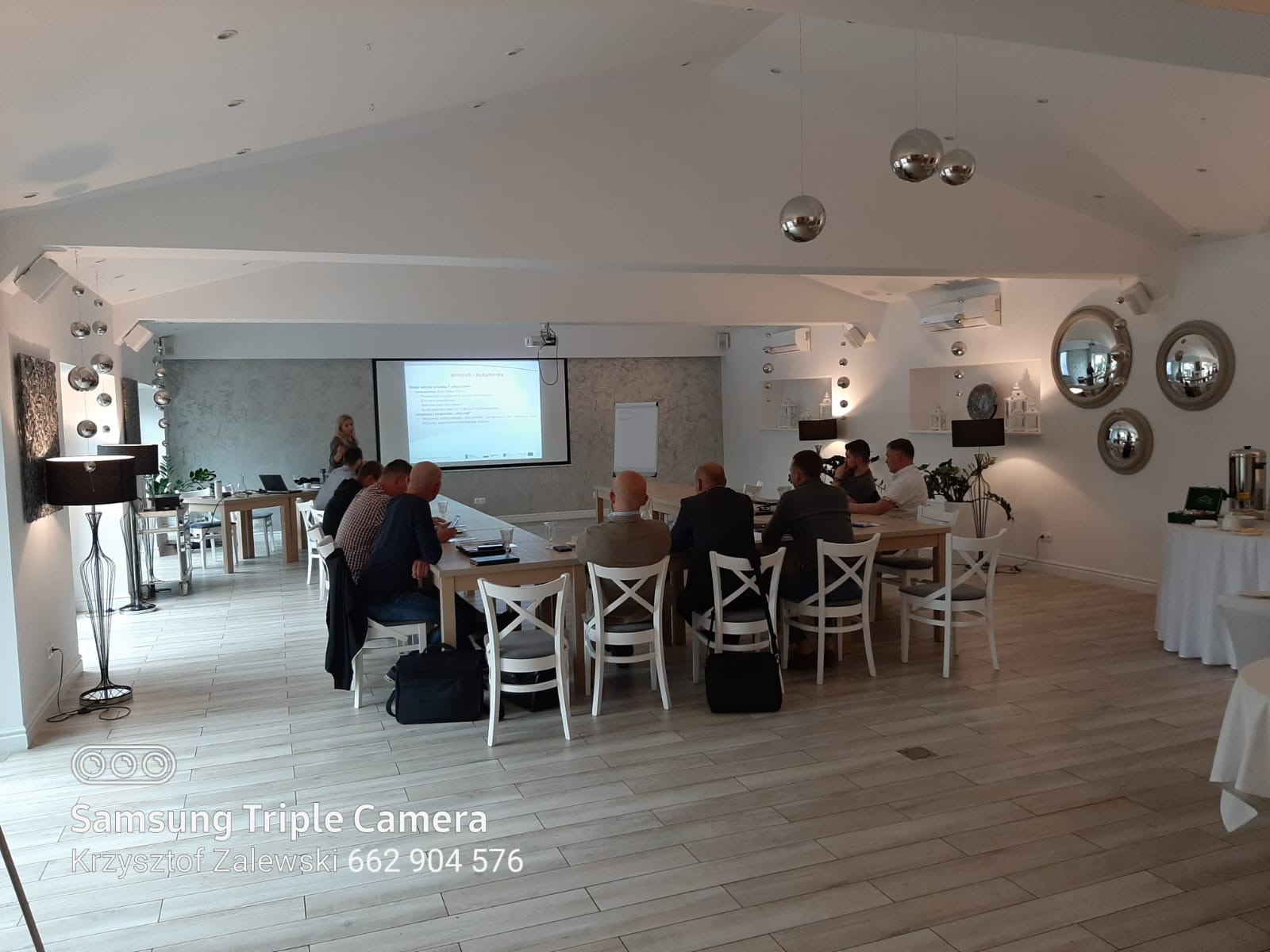 Sala konferencyjna z grupą osób siedzących przy stole i prezentacją wyświetlaną na ekranie, widoczne lampy wiszące w stylu disco, dekoracyjne lustra na ścianie i jasna podłoga imitująca drewno.