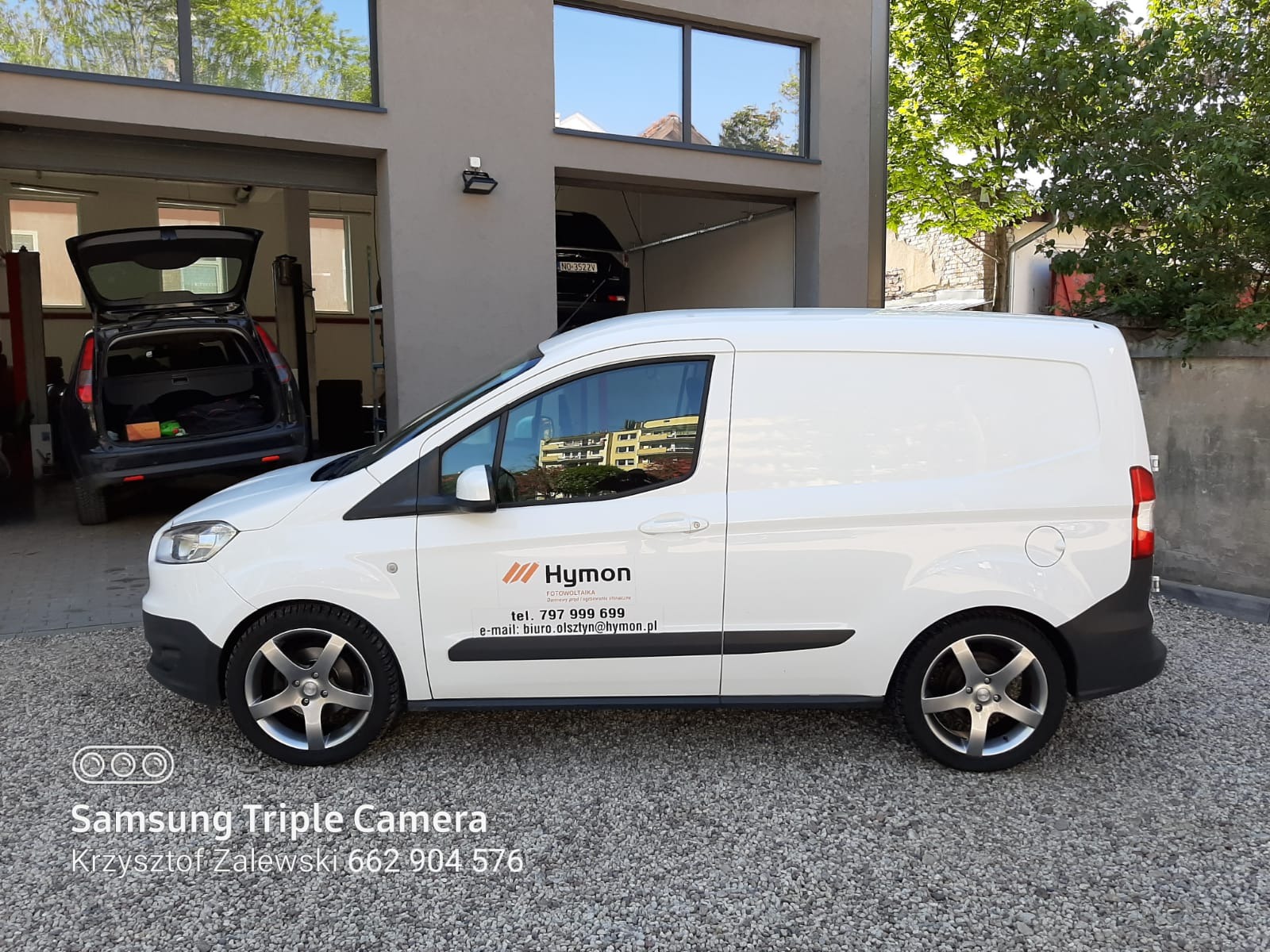 Biały samochód dostawczy firmy Hymon z logo i danymi kontaktowymi, zaparkowany na żwirowym podjeździe przed budynkiem z otwartym garażem, w którym stoi samochód osobowy.