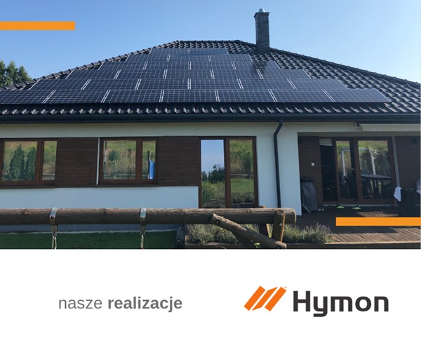 Panel słoneczny na dachu domu jednorodzinnego, widok z poziomu ziemi, w tle błękitne niebo i zieleń drzew, w dolnej części kadru widoczny drewniany płotek.