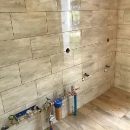 Ściana i podłoga wyłożone płytkami imitującymi drewno w łazience, widoczne przyłącza wodne i otwory w ścianie pod armaturę.