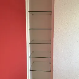 Wnęka ścienna z zamontowanymi szklanymi półkami, widoczna czerwona ściana obok i biała ściana z teksturą.