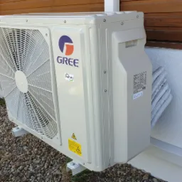 Zewnętrzna jednostka klimatyzacji Gree, zamontowana na niskim fundamencie z kamyków, z widocznymi przewodami chłodniczymi i tabliczką znamionową, przy białej ścianie budynku z drewnianą okładziną...