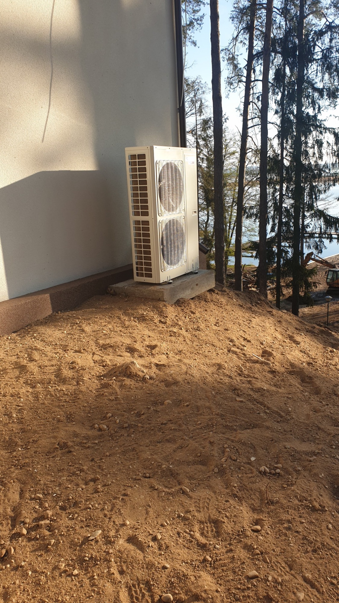 Zewnętrzna jednostka klimatyzacji, dwuwentylatorowa, zamontowana na betonowym fundamencie przy ścianie budynku na nierównym terenie, w tle drzewa i jezioro.