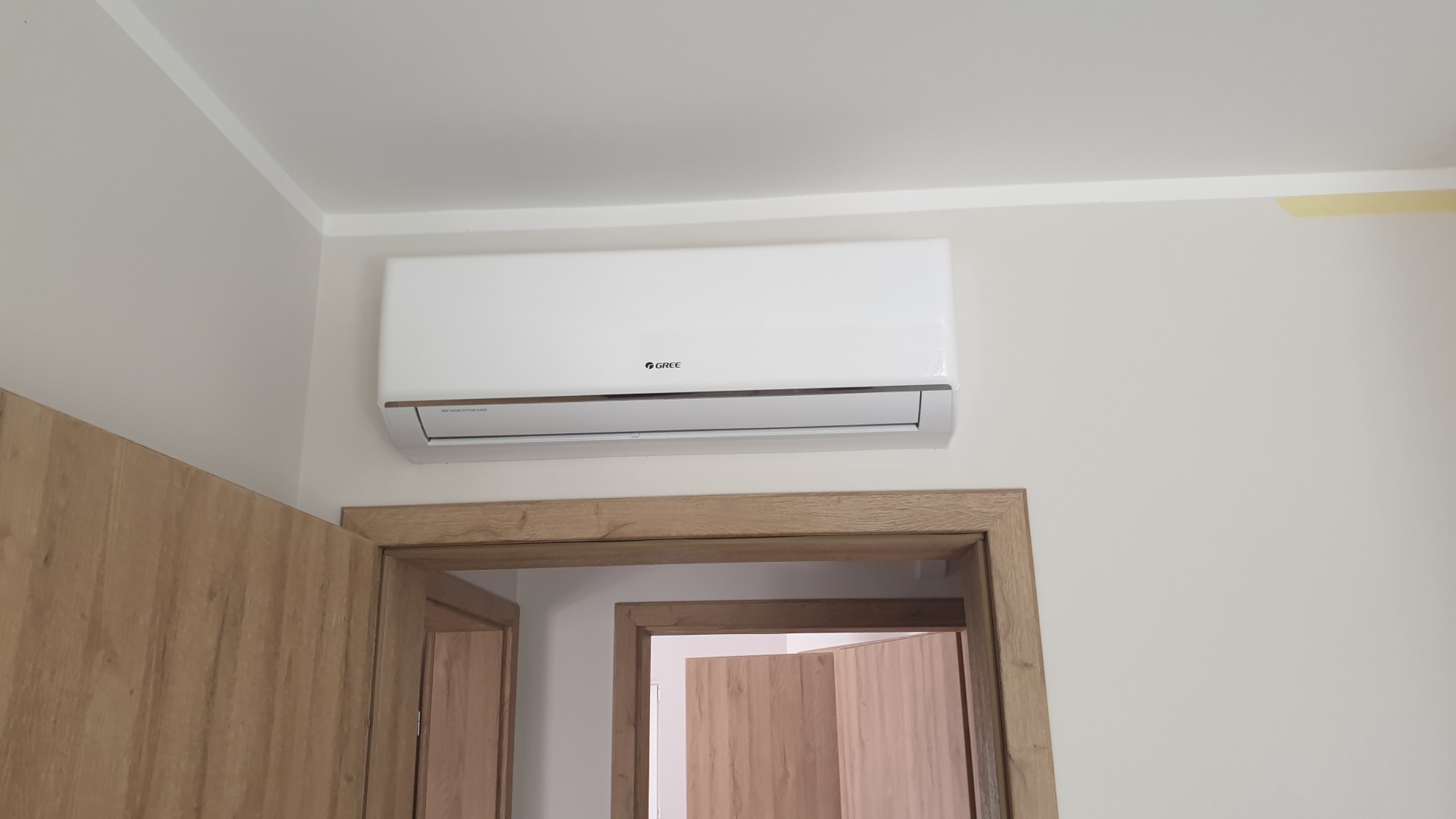 Biała jednostka wewnętrzna klimatyzatora marki Gree zamontowana na ścianie nad drewnianą futryną drzwiową, widok z dołu.