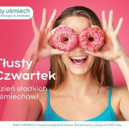 OBIEKTYWna Klementyna Sipko - Uśmiechnięta kobieta trzymająca przed oczami dwa pączki z różowym lukrem i posypką na tle różowego studia, z widocznym logo firmy stomatologicznej w lewym górnym rogu i hasłem reklamowym 'Tłusty...