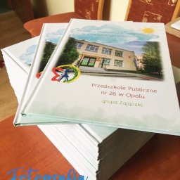 OBIEKTYWna Klementyna Sipko - Stos albumów pamiątkowych z ukończenia przedszkola, z fotografią budynku Przedszkola Publicznego nr 26 w Opolu i napisem 'Pamiątka ukończenia przedszkola w roku szkolnym 2019/2020'.