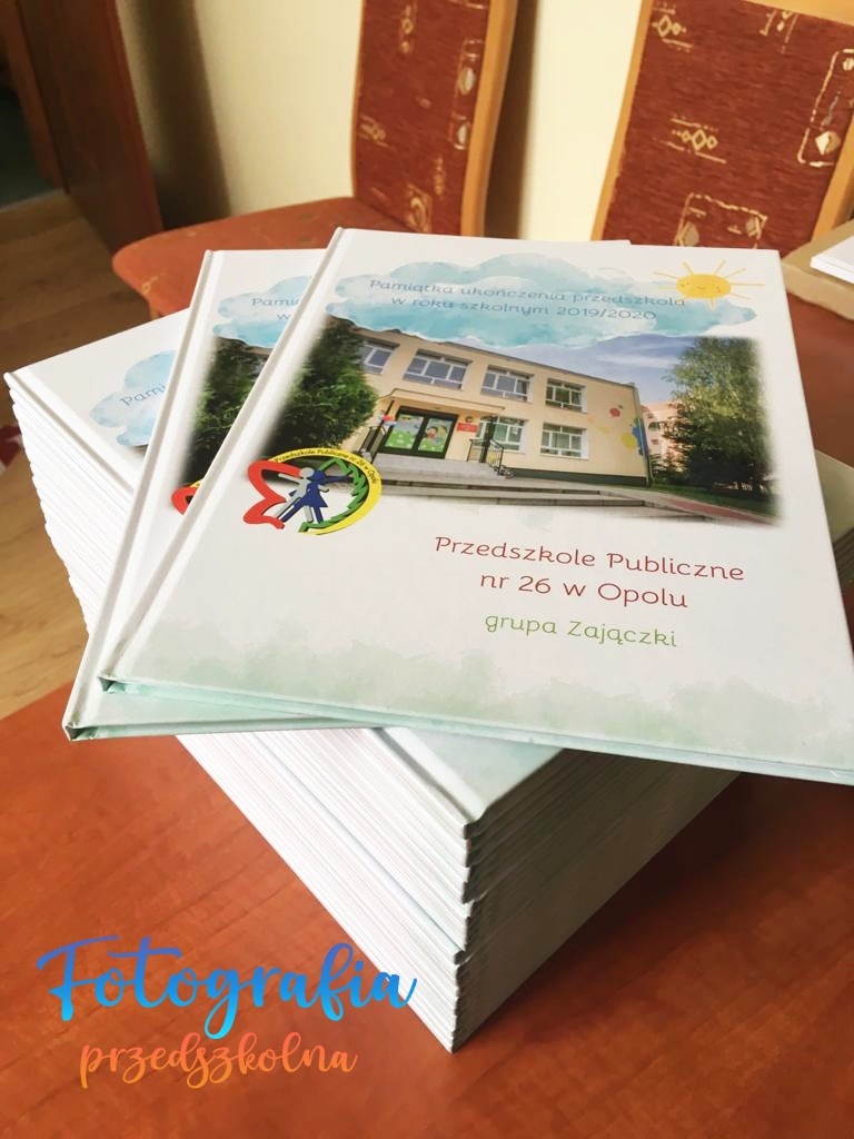 Stos albumów pamiątkowych z ukończenia przedszkola, z fotografią budynku Przedszkola Publicznego nr 26 w Opolu i napisem 'Pamiątka ukończenia przedszkola w roku szkolnym 2019/2020'.
