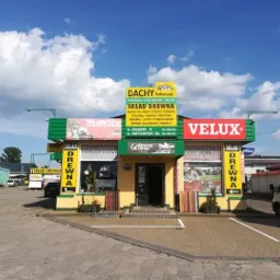 Fasada składu drewna i materiałów budowlanych z szyldami Dachy Kolberczyk, Velux, Makita, oferującego blachy dachowe, systemy rynnowe, stolarkę okienną i obróbki blacharskie, z ekspozycją paneli...