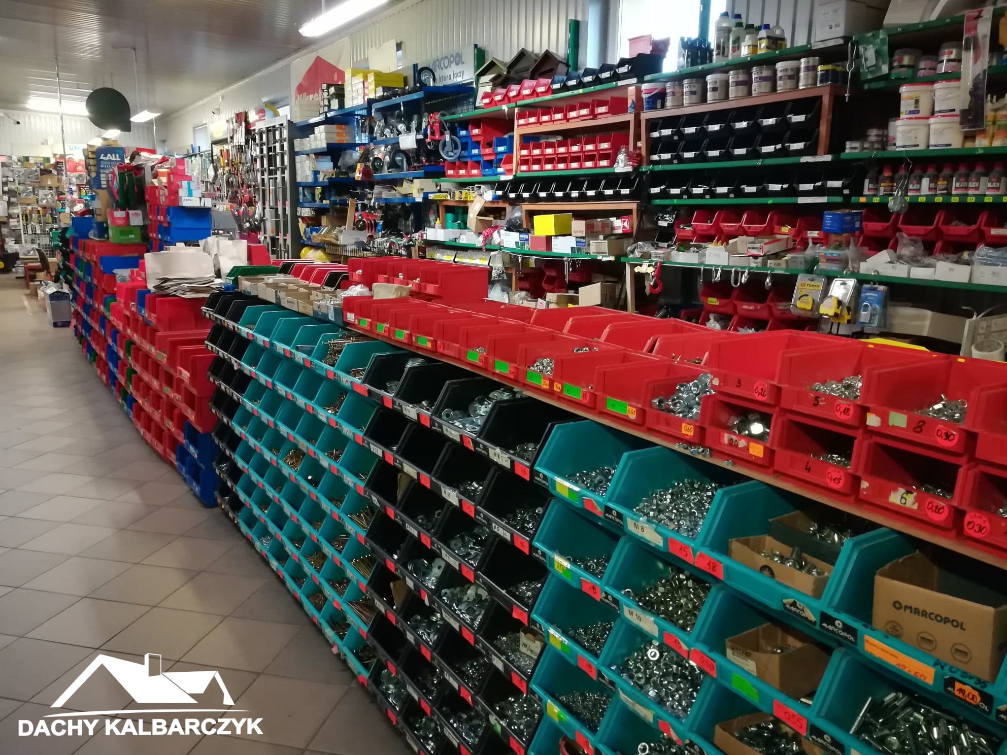 Wnętrze sklepu z materiałami budowlanymi, regały wypełnione różnymi rodzajami śrub, nakrętek i drobnych elementów metalowych w kolorowych pojemnikach, widoczne logo firmy 'Dachy Kalbarczyk'.