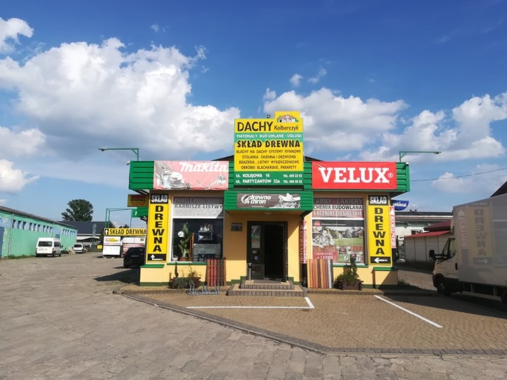 Fasada składu drewna i materiałów budowlanych z szyldami Dachy Kolberczyk, Velux, Makita, oferującego blachy dachowe, systemy rynnowe, stolarkę okienną i obróbki blacharskie, z ekspozycją paneli...