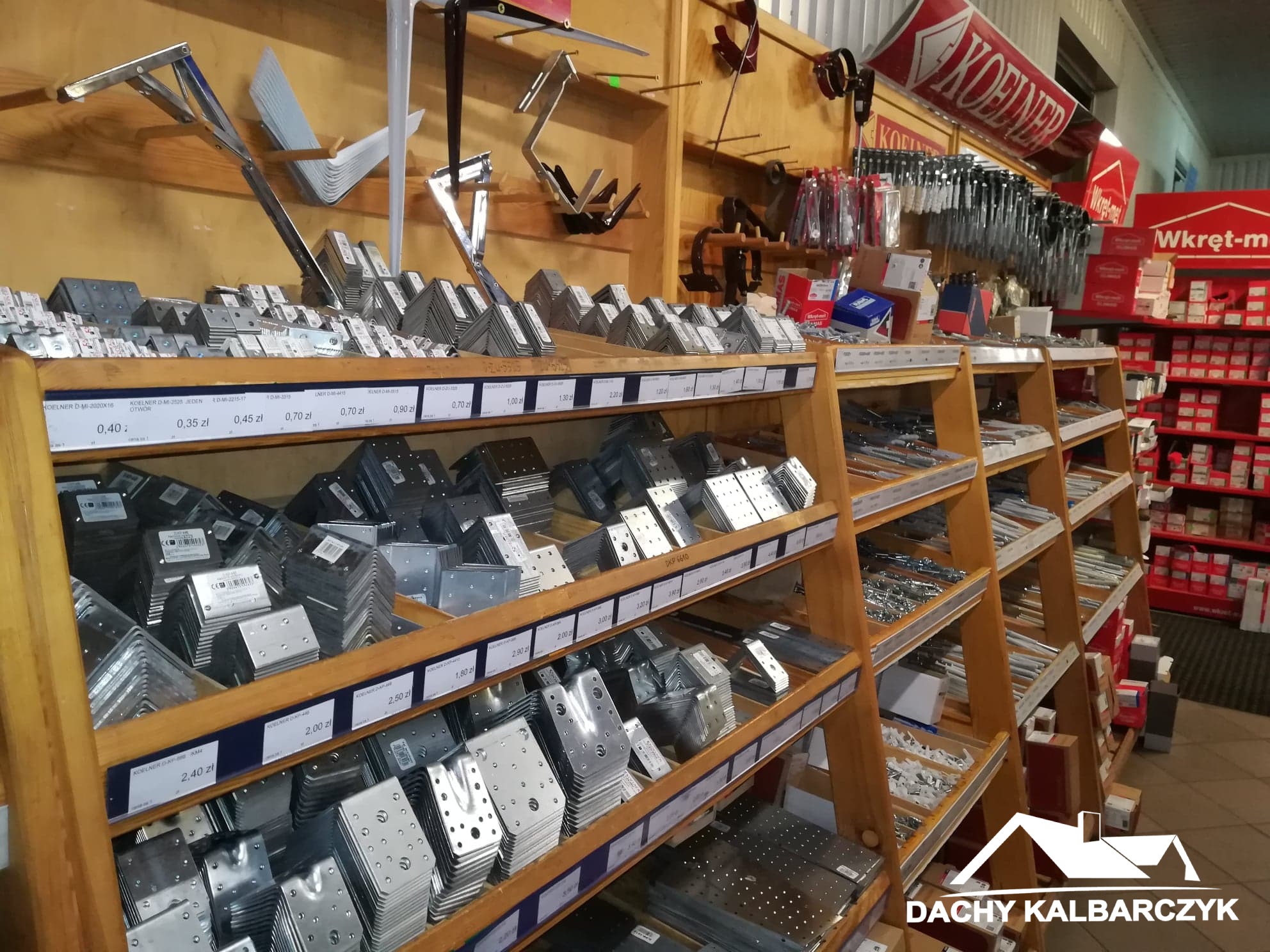 Sklep budowlany: półki z metalowymi łącznikami kątowymi, śrubami i akcesoriami do konstrukcji dachowych, widoczne ceny na etykietach.