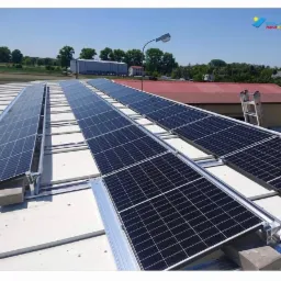 Instalacja paneli słonecznych na płaskim dachu, widoczne betonowe bloczki jako obciążenie, drabina oparta o budynek w tle, logo firmy w prawym górnym rogu.