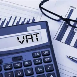 Kalkulator z wyświetlanym napisem VAT, obok wykres słupkowy, okulary i długopis na jasnym tle.