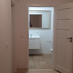 Bud-mar Mariusz Kos usługi ogólnobudowlane. - Widok z korytarza na wykończoną, minimalistyczną łazienkę z drewnopodobną podłogą, białymi ścianami, wiszącą szafką z umywalką i dużym lustrem w srebrnej ramie.