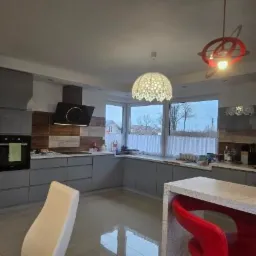 Nowoczesna kuchnia z szarymi szafkami, drewnianymi akcentami na ścianie, wyspą z czerwonymi hokerami i designerskimi lampami wiszącymi.