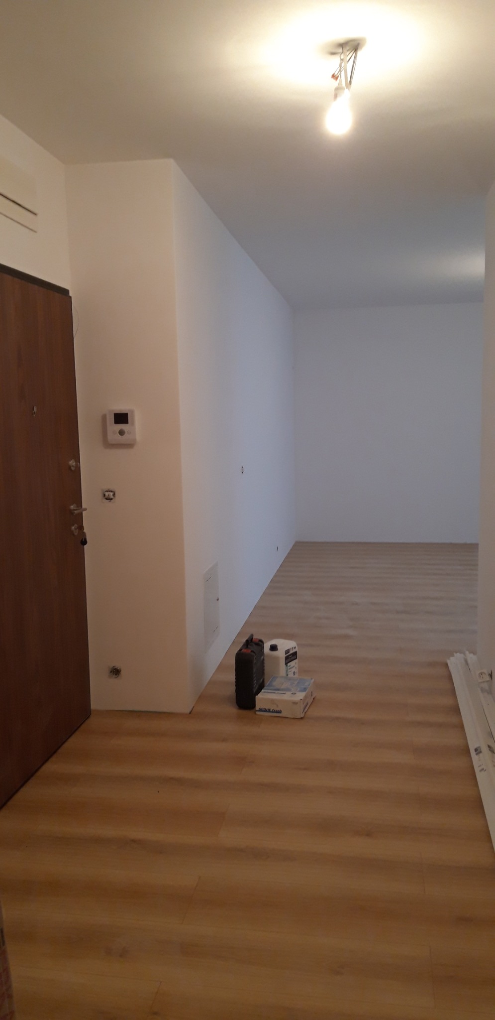 Nowe panele podłogowe w jasnym odcieniu drewna w przedpokoju, przy ścianie stoją materiały budowlane: wiadro, pojemnik i karton.