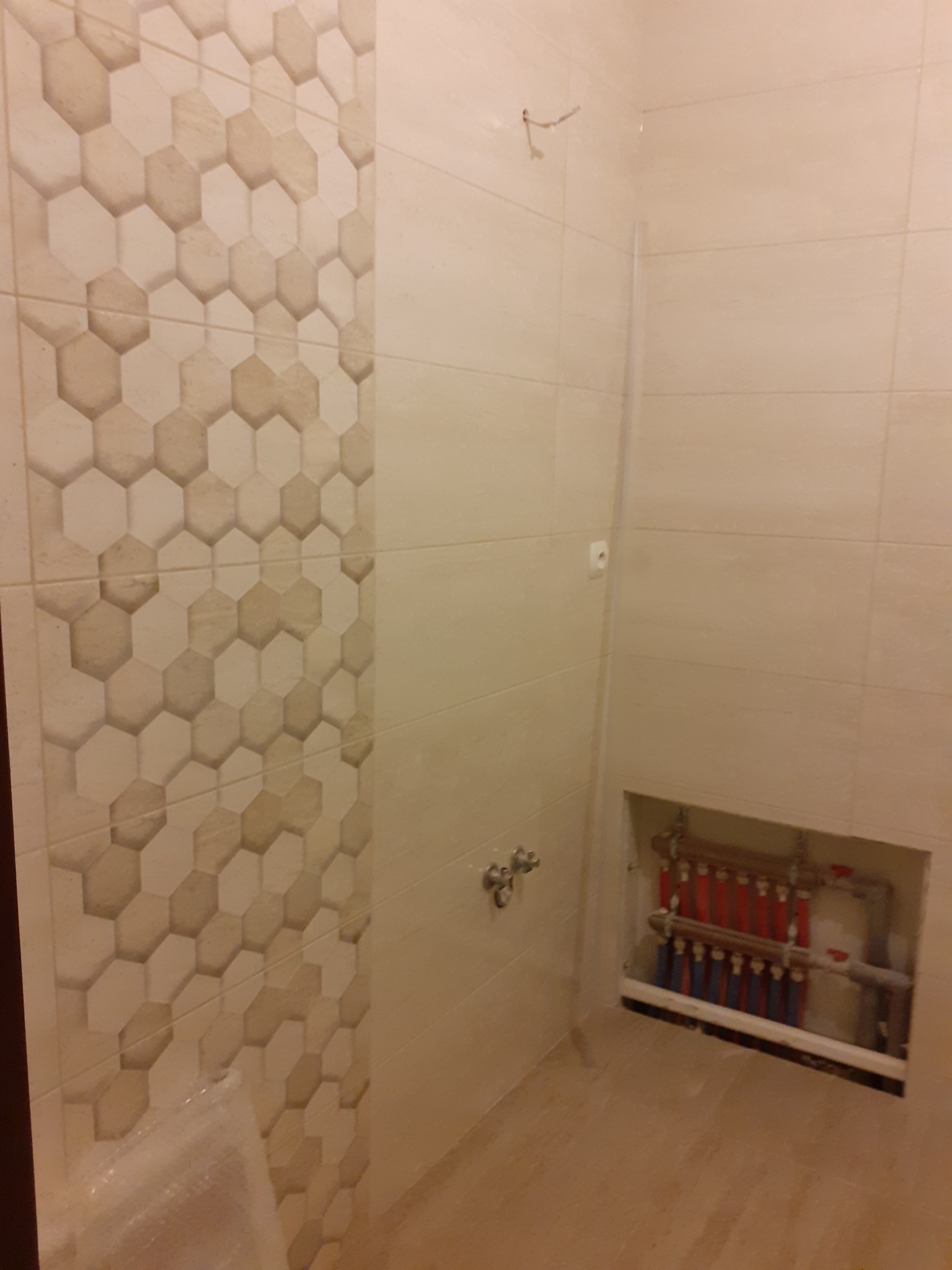 Jasna łazienka w trakcie remontu, z dekoracyjnymi heksagonalnymi płytkami na ścianie i odkrytą instalacją hydrauliczną w niszy.