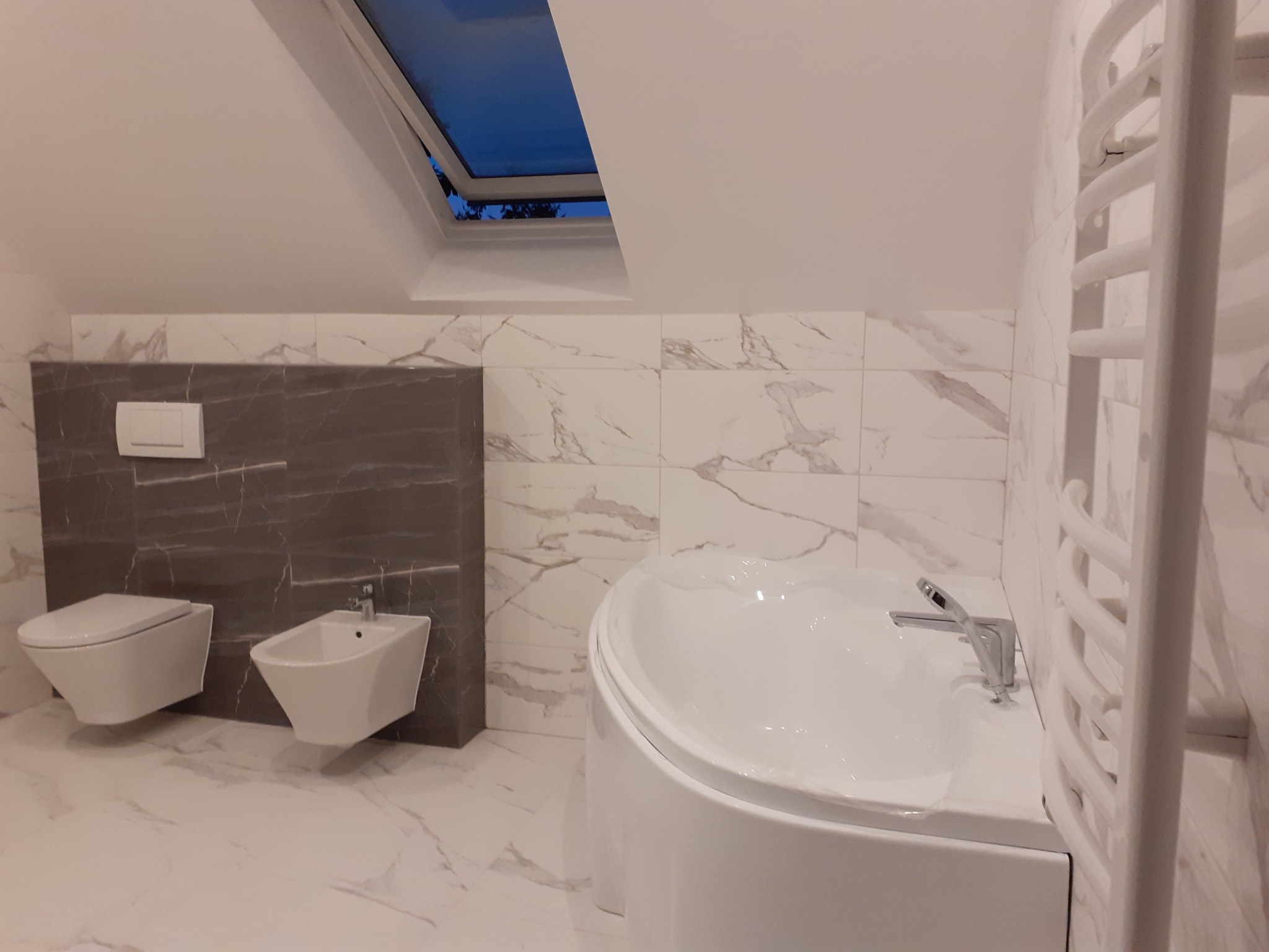 Wykończona łazienka na poddaszu z oknem dachowym, białą armaturą (toaleta, bidet, wanna) i grzejnikiem drabinkowym, płytki imitujące marmur.
