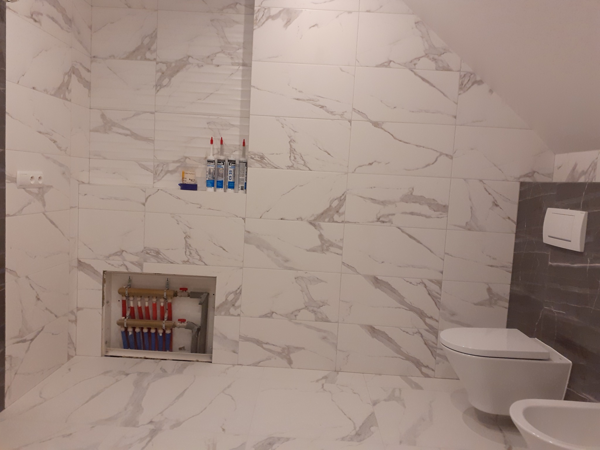 Wykończona łazienka z białymi płytkami imitującymi marmur, wisząca toaleta i bidet, oraz rozdzielacz ogrzewania podłogowego w zabudowie.