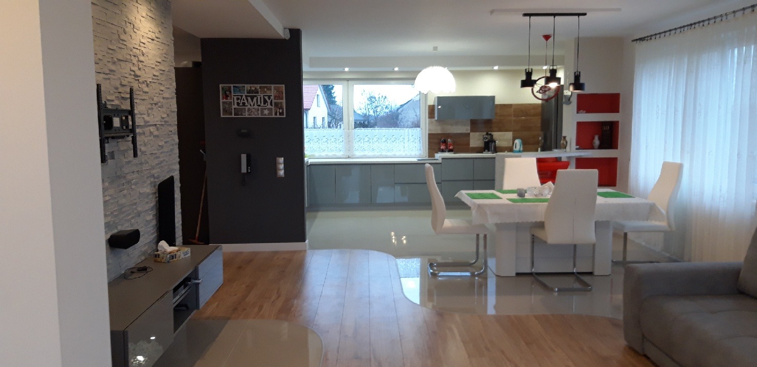 Jasne, przestronne wnętrze domu z widokiem na kuchnię z szarymi szafkami, jadalnię z białym stołem i krzesłami oraz salon z szarą sofą. Na ścianie w salonie dekoracyjna okładzina kamienna...