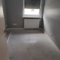 Pomalowane na szaro puste pomieszczenie z marmurową podłogą, białymi listwami przypodłogowymi i grzejnikiem pod oknem z roletami.