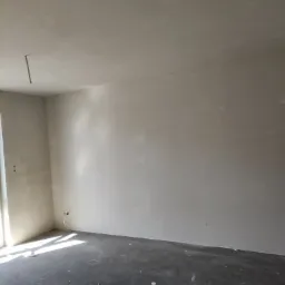 Pokój po nałożeniu gładzi gipsowej na ścianach i suficie, widoczne wystające kable elektryczne i fragment okna z wpadającym światłem.