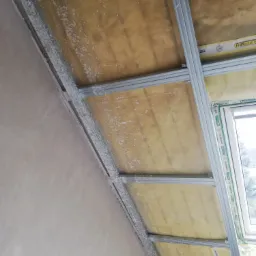 Konstrukcja metalowa sufitu podwieszanego z izolacją z wełny mineralnej, widoczne ślady szpachlowania na profilach i ścianie, okno w tle.