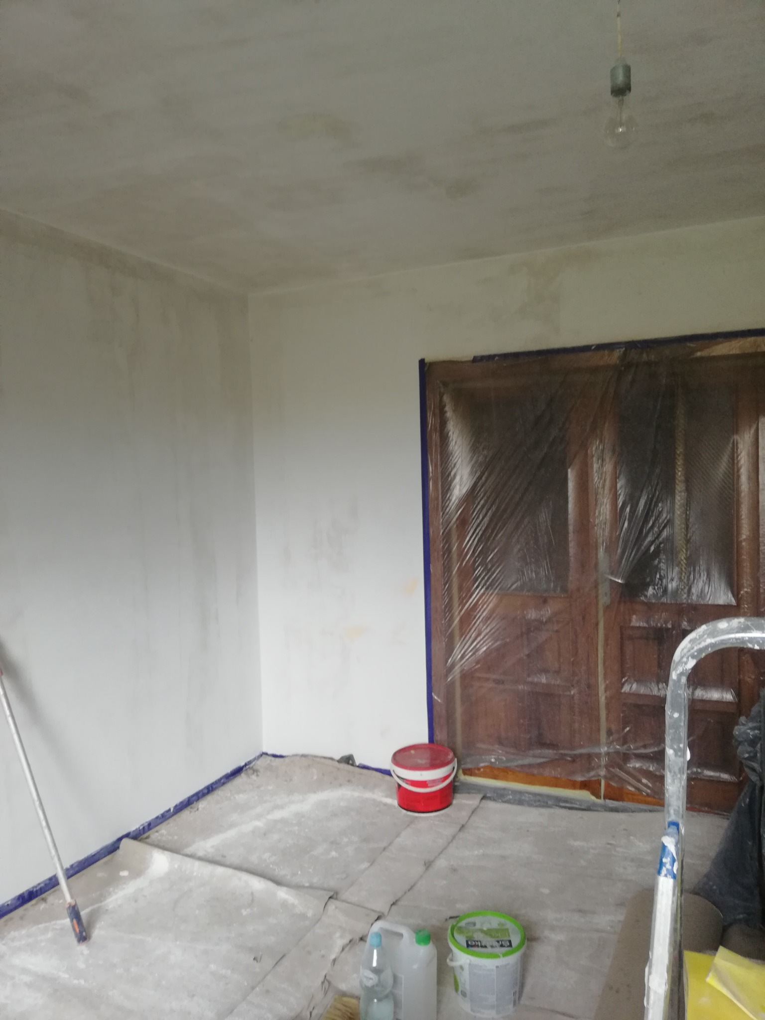 Pokój w trakcie przygotowań do malowania: zabezpieczone folią drzwi, podłoga przykryta kartonem, widoczny wałek malarski i pojemniki z farbą.