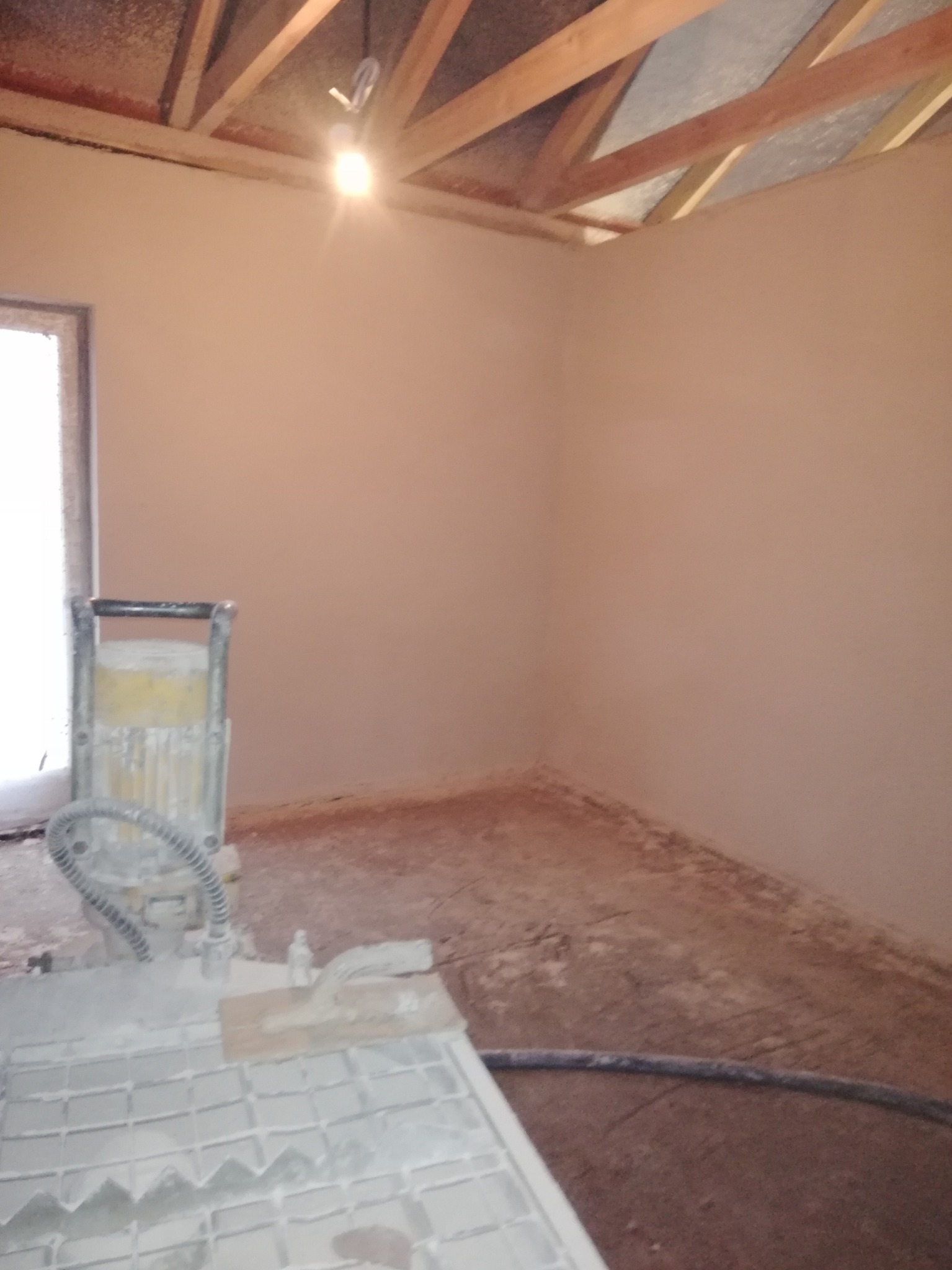 Wnętrze pokoju w trakcie prac wykończeniowych, ściany pokryte świeżym tynkiem, widoczna maszyna do gładzi i surowy drewniany strop.