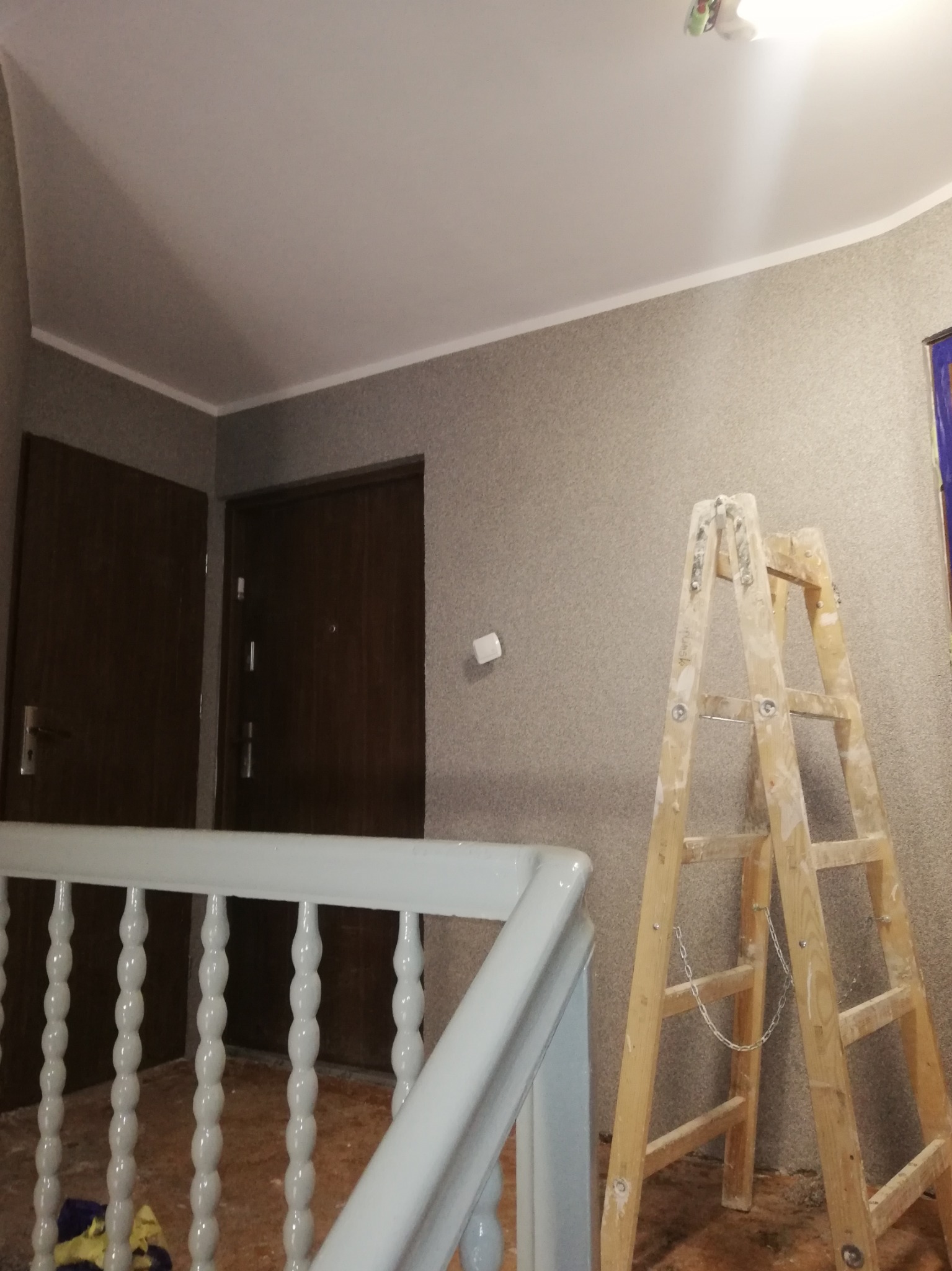 Klatka schodowa w trakcie odświeżania: jasna balustrada, drewniana drabina oparta o szarą ścianę z widocznymi śladami farby, dwie ciemne drzwi w tle.