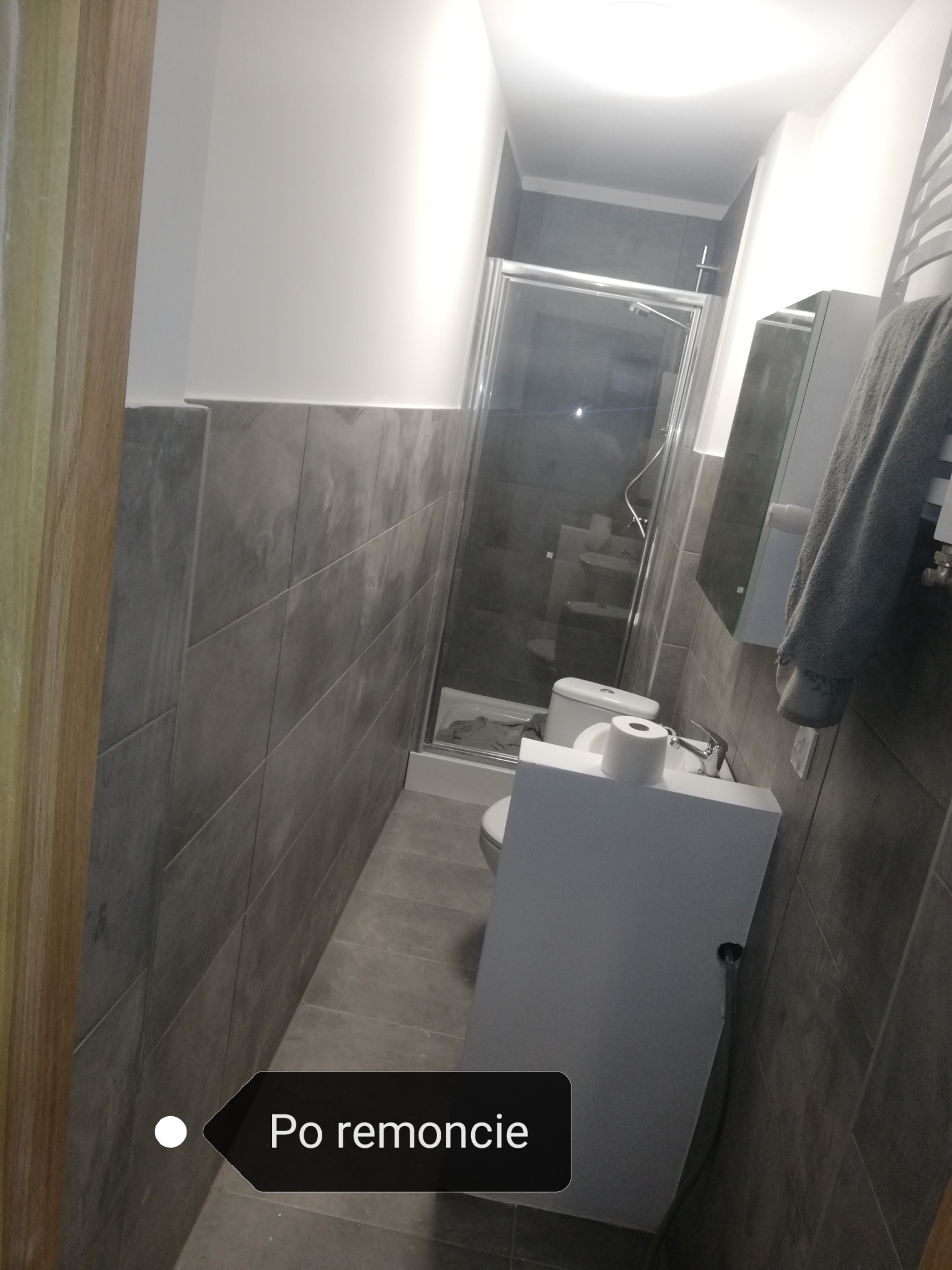 Wąska łazienka po remoncie z szarymi płytkami, kabiną prysznicową i podwieszaną toaletą z podajnikiem na papier toaletowy.