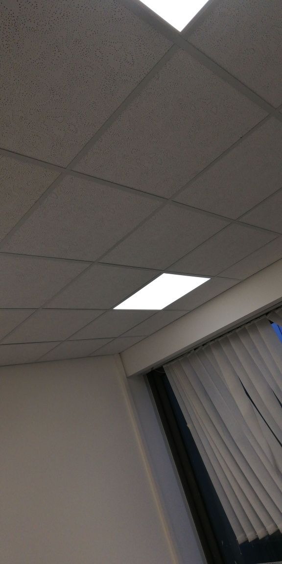 Kasetonowy sufit w biurze z oświetleniem LED i fragmentem okna z żaluzjami w tle.